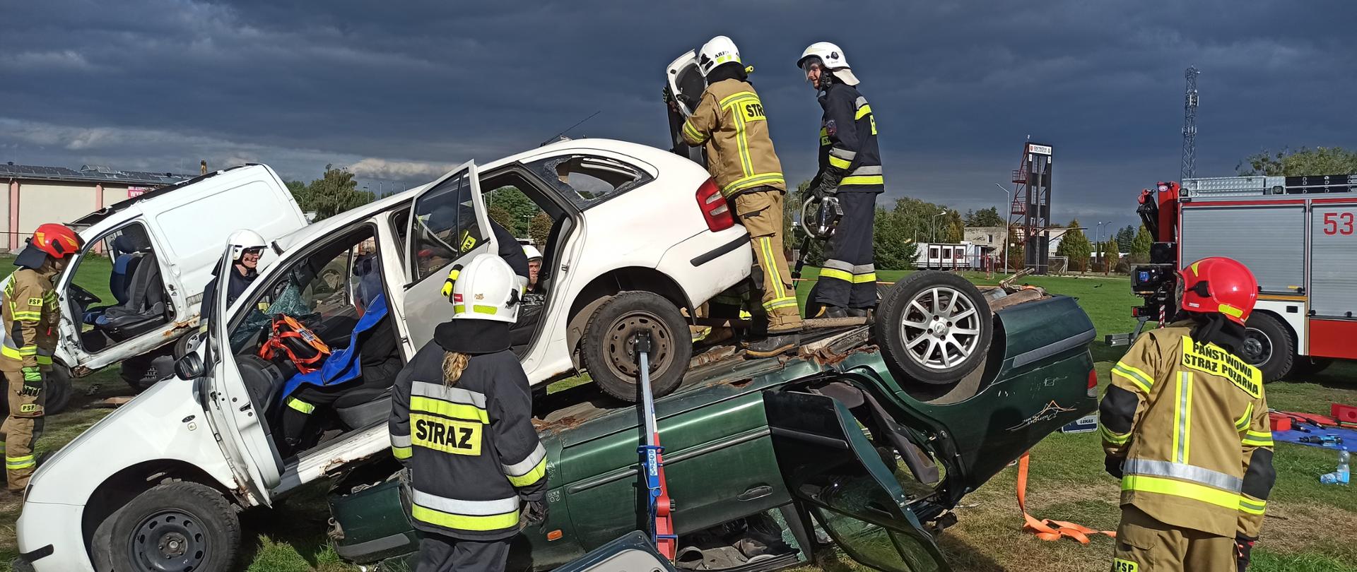 Strażacy w umundurowaniu specjalnym i hełmach ćwiczący ratownictwo techniczne na wrakach samochodów, w tle pojazdy pożarnicze