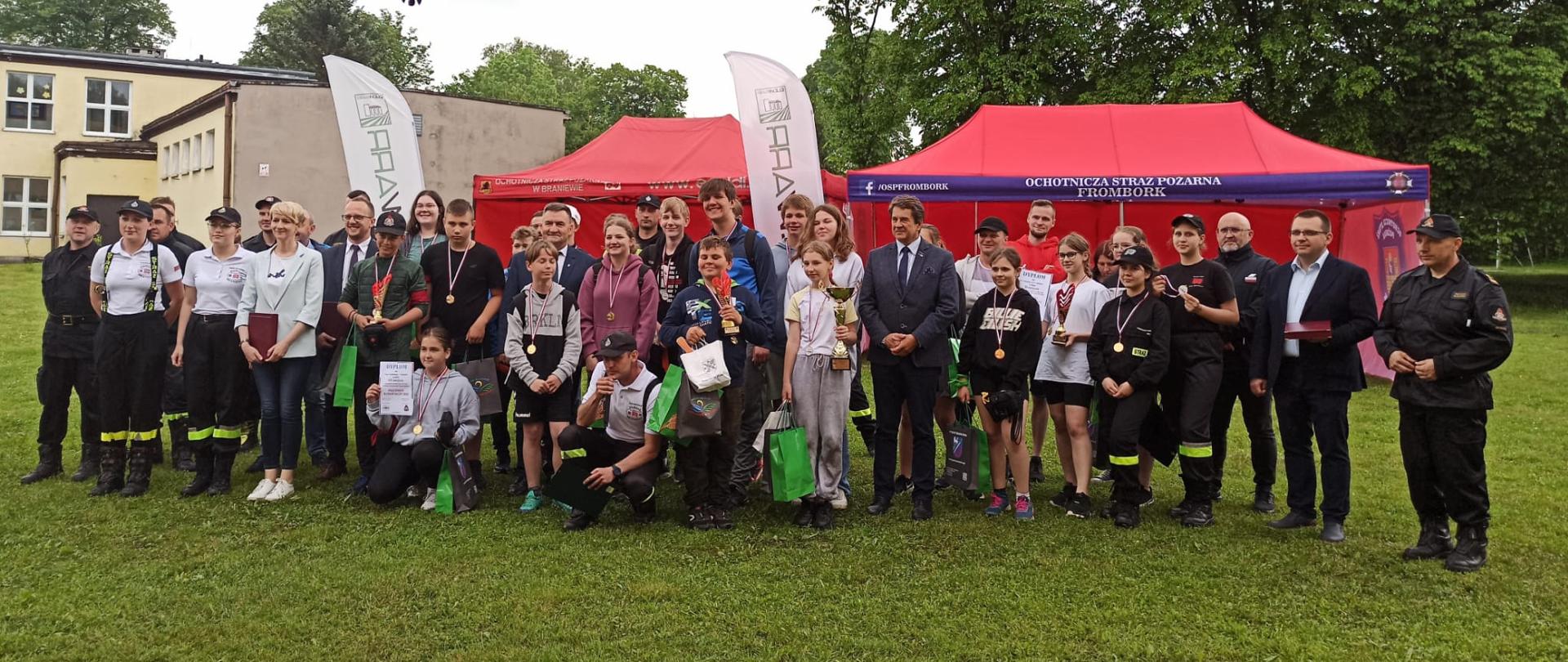 Na zdjęciu duża grupa uczestników i organizatorów zawodów ustawieni w szeregu na zielonej trawie boiska. Za nimi czerwone namioty i drzewa.