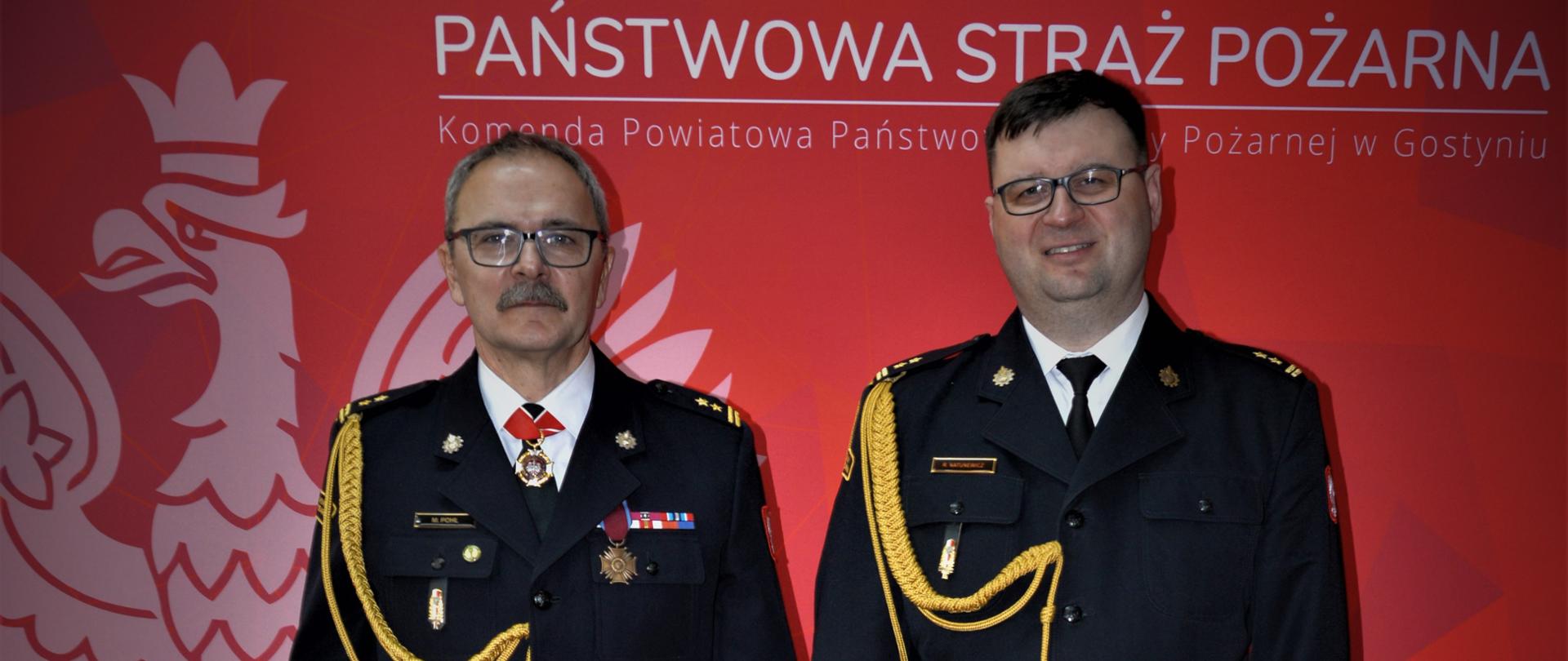 dwóch strażaków stoi obok siebie, w tle czerwona ścianka z napisem państwowa straż pożarna