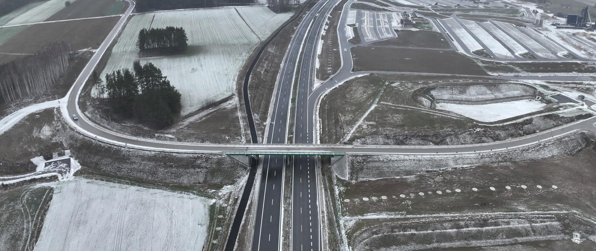 Widok z lotu ptaka na dwujezdniową drogę ekspresową z wiaduktem nad jezdnią, otoczoną polami częściowo pokrytymi śniegiem. Krajobraz zimowy, z szarym, pochmurnym niebem w tle i rozległymi terenami rolniczymi po obu stronach drogi.