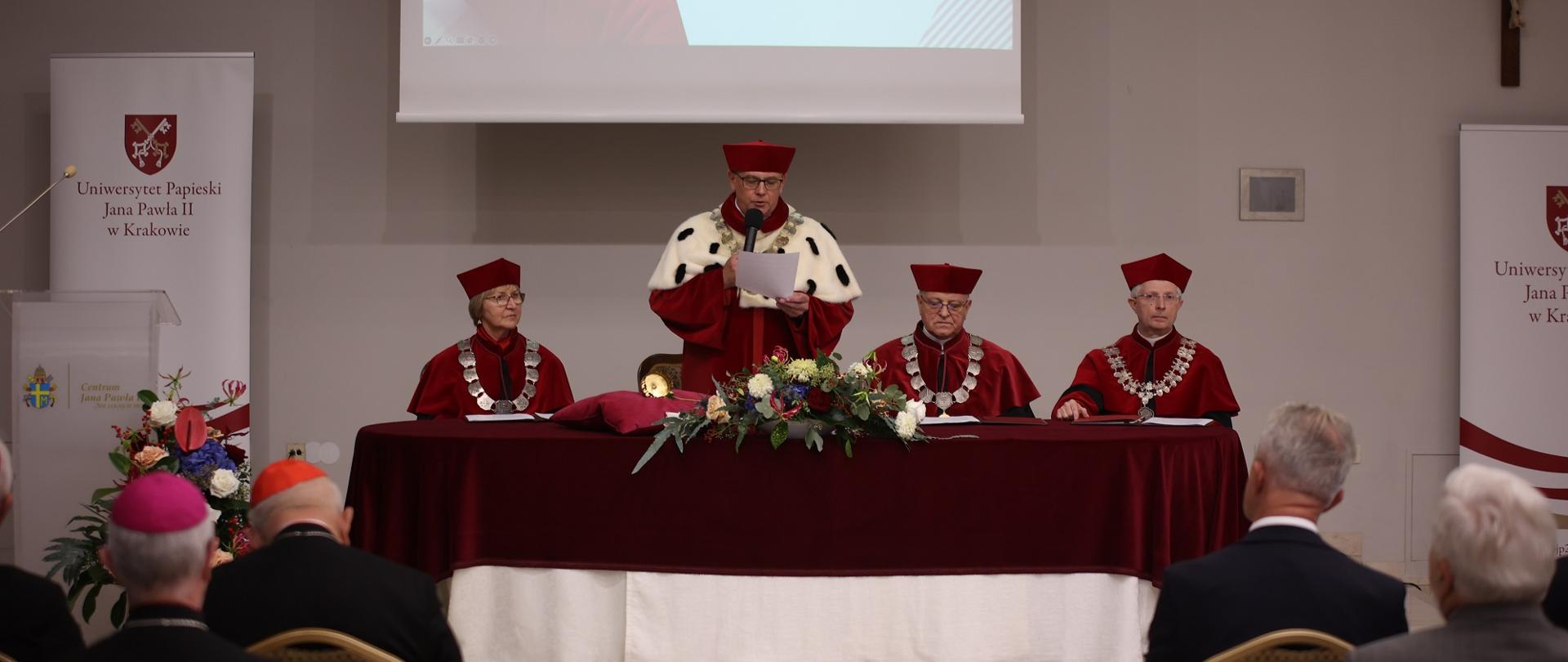 Widok od tyłu na salę, z przodu przy nakrytym czerwonym materiałem stole siedzą trzy osoby w czerwonych ceremonialnych strojach, pośrodku stoi mężczyzna w takim samym stroju z gronostajowym kołnierzem i mówi do trzymanego w ręku mikrofonu.