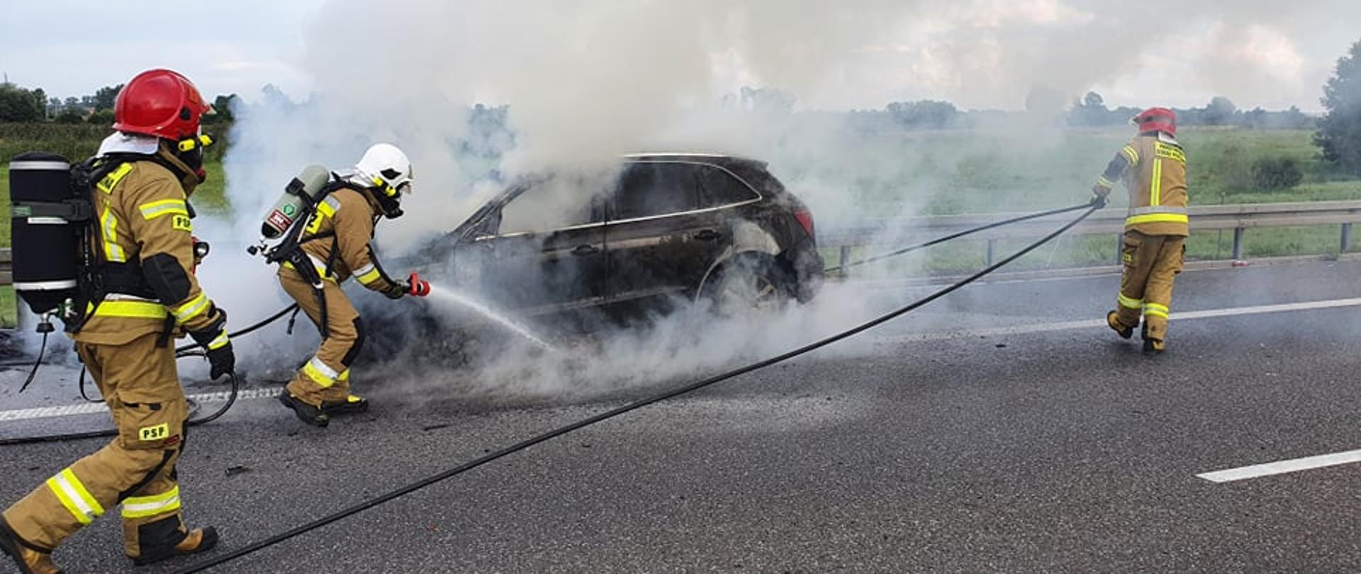 Pożar auta na S5. Jezdnia trasy S5 w kierunku Poznania. Na pasie awaryjnym stoi palący się samochód osobowy. Wokół niego zadymienie. Trzech strażaków prowadzi akcję gaśniczą. W tle drzewa.