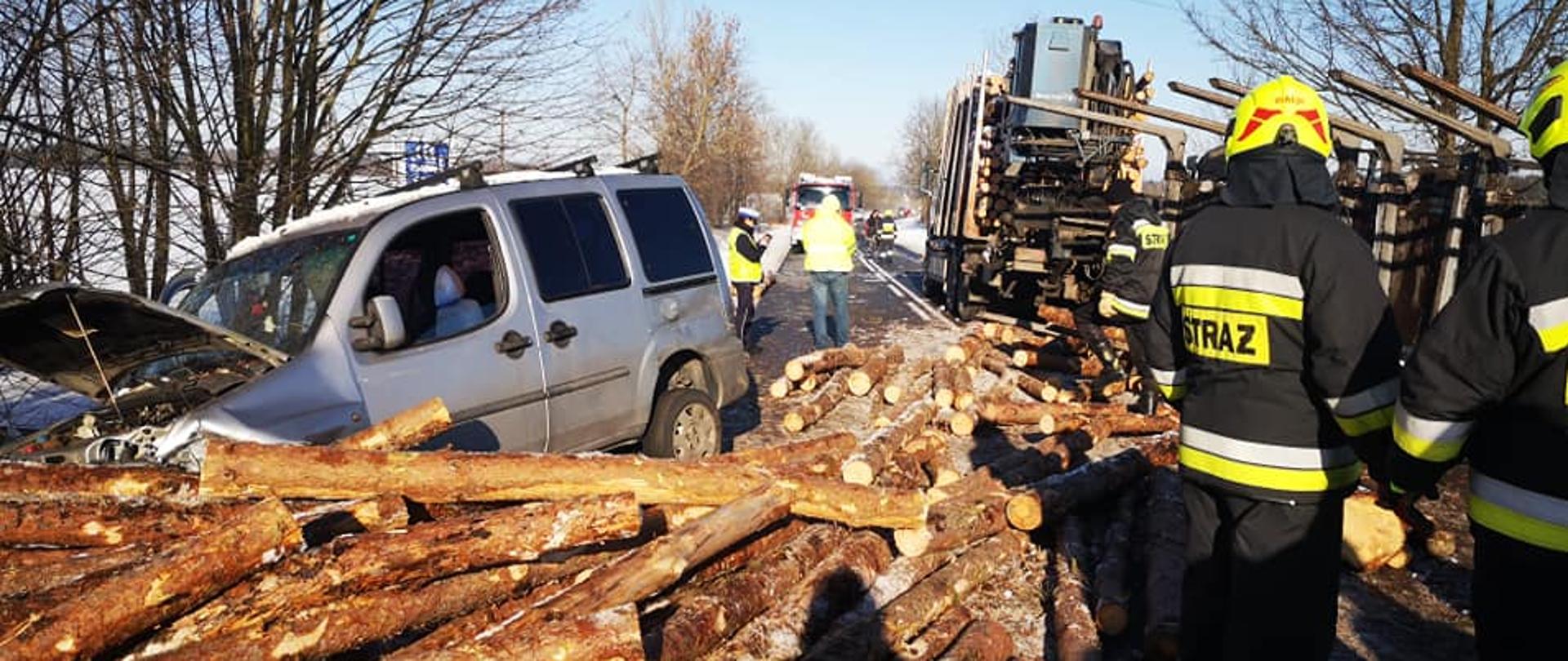 Zdjęcie przedstawia drogę pokrytą delikatnie śniegiem na którą są wysypane kłody drewna sosnowego. Po lewej stronie widzimy srebrny samochód dostawczy z podniesiona maską, za autem widzimy dwóch policjantów. Po prawej stronie widzimy samochód ciężarowy oraz przewróconą przyczepę, która przewoziła kłody. Przed przyczepą znajdują się trzech strażaków w czarnych ubraniach specjalnych oraz żółtych hełmach. W tle widać pole, niebo, drzewa. Zdjęcie zrobione za dnia.