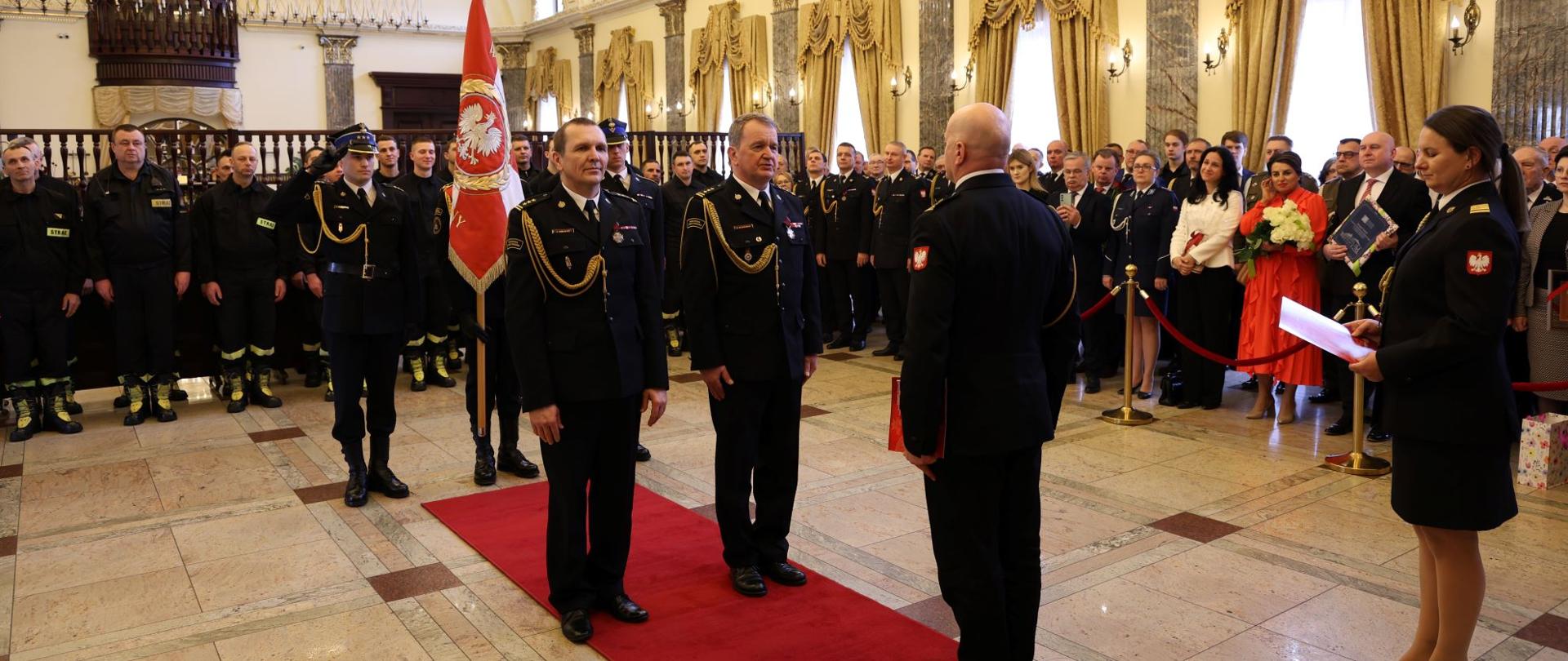 Na czerwonym dywanie stoi dwóch strażaków, trzeci przed nimi, zwrócony przodem do tamtych. Za nimi trzech innych ze sztandarem. W dai widać kilkadziesiśt osób, gości uroczystości. Z prawej strony strażak kobieta, trzyma dokumenty.