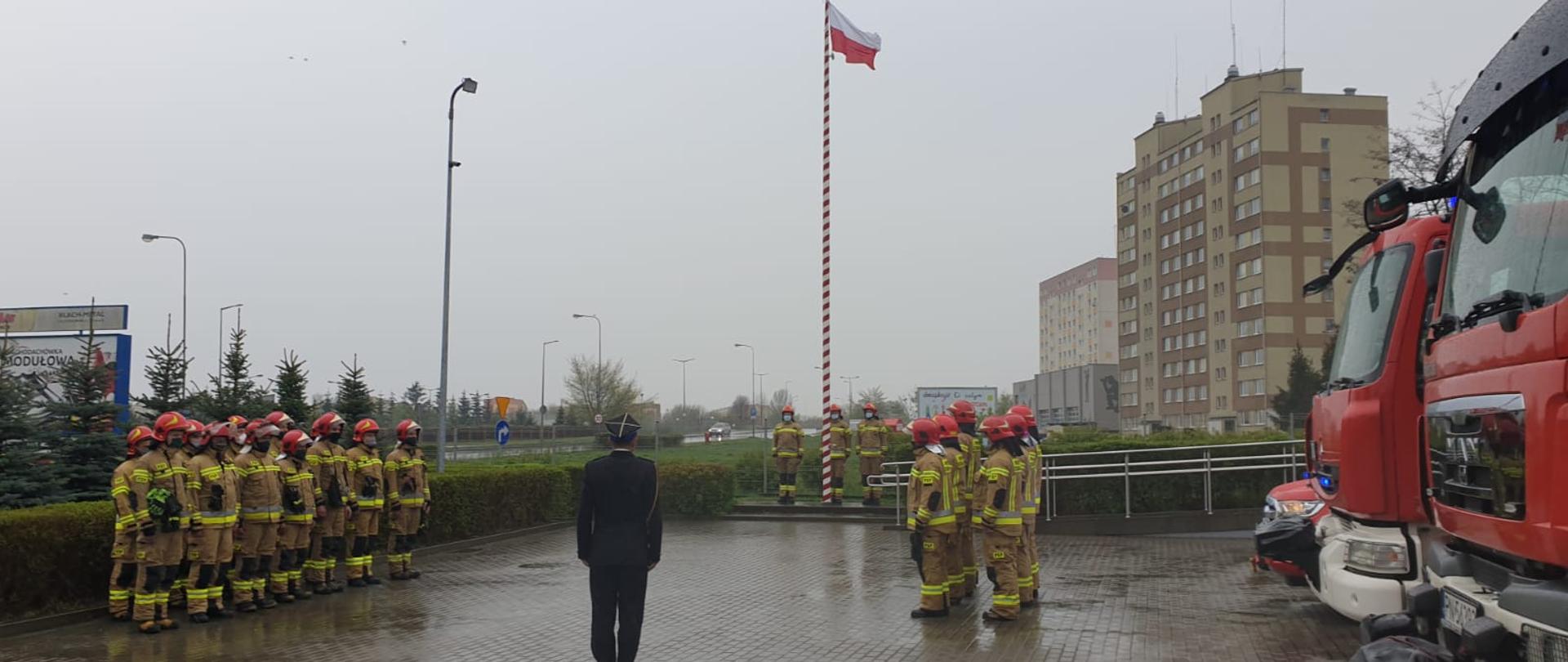 Zdjęcie przedstawia strażaków podczas uroczystej zbiórki z okazji Dnia Flagi. Na zdjęciu stoją strażacy na placu przed strażnicą oraz widać podniesioną flagę i samochody strażackie