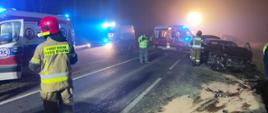 Na zdjęciu na silnie oświetlonej przez pojazdy drodze widoczne trzy pojazdy pogotowia ratunkowego. Na poboczu czarny rozbity samochód osobowy, przed którym rozsypane są części pojazdu. Obok uszkodzonego pojazdu widać plecy przechodzącego strażaka. Na pierwszym planie po lewej stronie widać plecy strażaka z włączoną latarką umieszczoną przy czerwonym hełmie zwrócony twarzą w lewą stronę. W centralnej części zdjęcia mężczyzna w jasnozielonej kamizelce i w maseczce na twarzy rozmawiający przez telefon komórkowy.