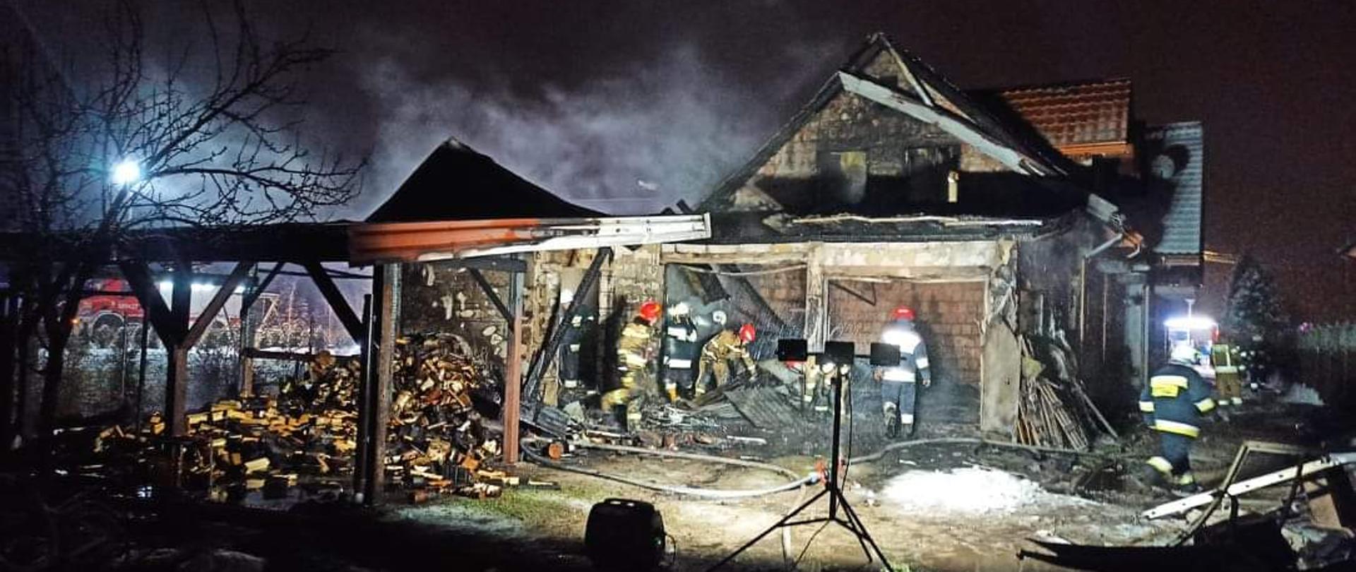 Na środku zdjęcia znajduje się budynek mieszkalny wniesiony z czerwonej cegły. Od przedniej strony, do budynku przylega murowany garaż wraz z zadaszoną wiatą, której konstrukcję tworzą drewniane słupy. Front budynku jest okopcony na czarno, w dwóch otworach okiennych nie ma okien. Nad budynkiem unosi biało – szary dym. Konstrukcja dachu garażu jest zniszczona, cześć pokrycia dachu znajduje się wewnątrz garażu. Do garażu prowadzą dwa duże otwory. Wewnątrz garażu znajduje się siedmioro strażaków – trzech w ubraniach specjalnych koloru piaskowego oraz czterech w ubraniach specjalnych koloru czarnego. Pod zadaszoną wiatą znajduje rozrzucone drewno, które było magazynowane na opał. Na pierwszym planie zdjęcia można zauważyć rozstawioną lampę z trzema halogenami, która oświetla miejsce akcji. Obok lampy, znajduje się agregat prądotwórczy. Przed garażem, na nieutwardzonym placu rozwinięte są węże strażackie. Widoczny jest śnieg oraz kałuże wody. W oddali po lewej stronie, można zauważyć samochód pożarniczy, który oświetla miejsce zdarzenia z wysokiego masztu. 