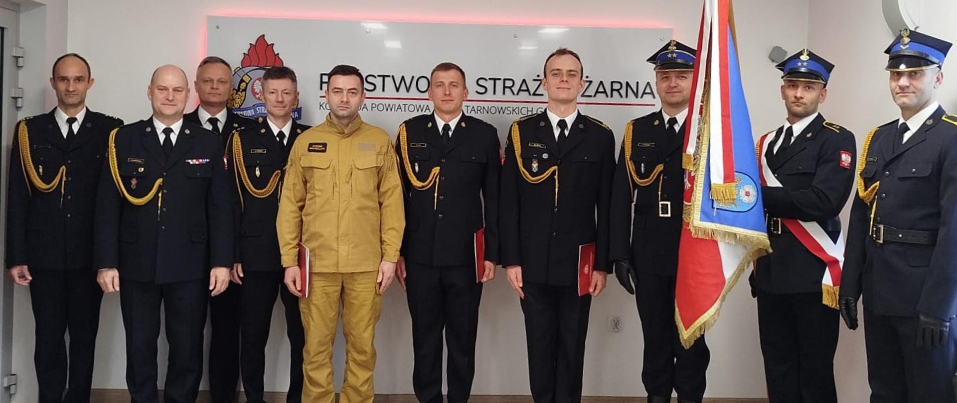 Na zdjęciu widać grupę dziesięciu mężczyzn w mundurach, ustawionych do pamiątkowej fotografii, po uroczystości związanej z Państwową Strażą Pożarną.
W centrum stoi mężczyzna w jasnym, beżowo-khaki mundurze, trzymający czerwoną teczkę w ręku. Pozostali mężczyźni są ubrani w ciemne mundury galowe PSP. Większość z nich ma na mundurach ozdobne złote sznury (szarfy), co jest typowe dla galowego umundurowania PSP. Trzech mężczyzn po prawej stronie stoi przy sztandarze i jest to poczet sztandarowy. Dwaj z nich mają na głowach czapki rogatywki galowe. Jeden z nich (trzymający drzewce sztandaru) ma na sobie biało-czerwoną szarfę. Sztandar: Po prawej stronie widać sztandar Komendy Powiatowej Państwowej Straży Pożarnej w Tarnowskich Górach, który jest częścią pocztu sztandarowego.
Tło: Na ścianie za grupą widnieje napis: "PAŃSTWOWA STRAŻ POŻARNA KOMENDA POWIATOWA PSP W TARNOWSKICH GÓRACH".