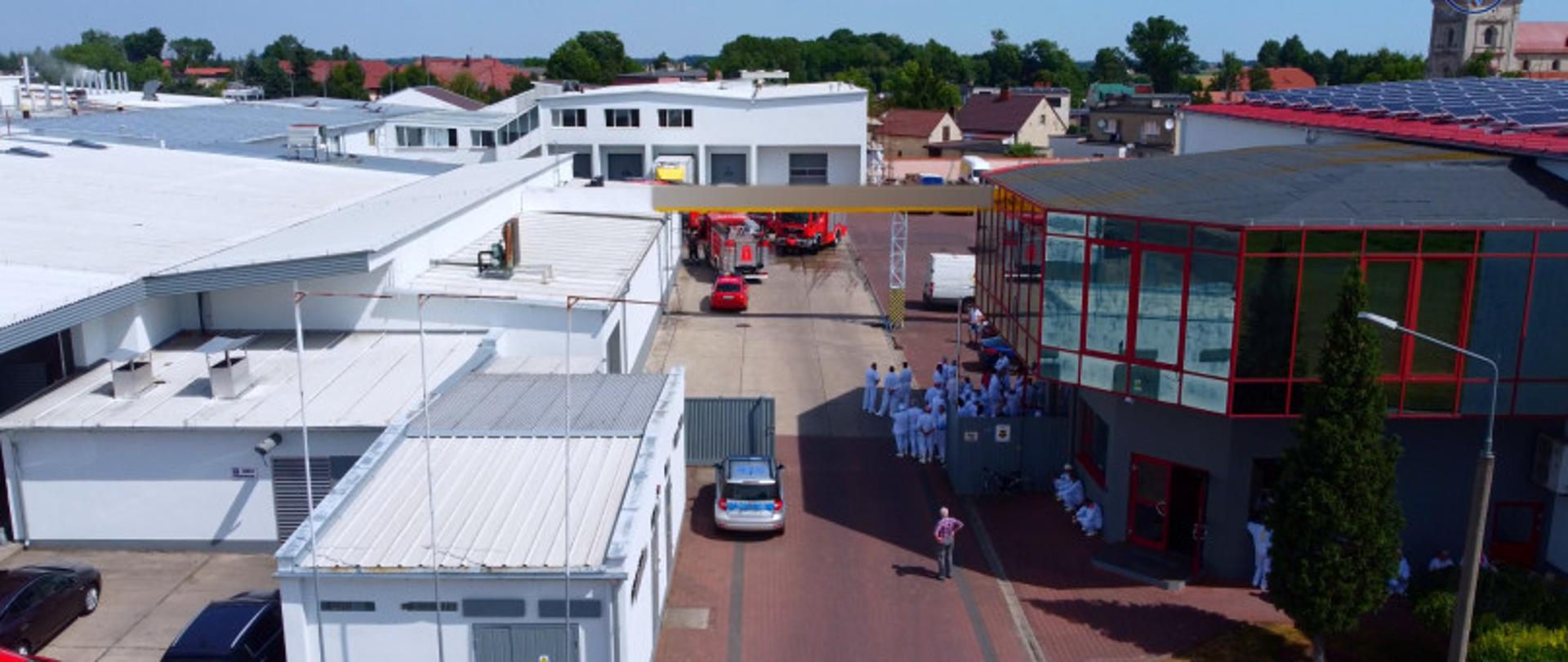 Zdjęcia przedstawia widok z lotu ptaka. Przedstawia ono zastępy straży pożarnej na placu zakładu produkcyjnego oraz ewakuowanych pracowników.
