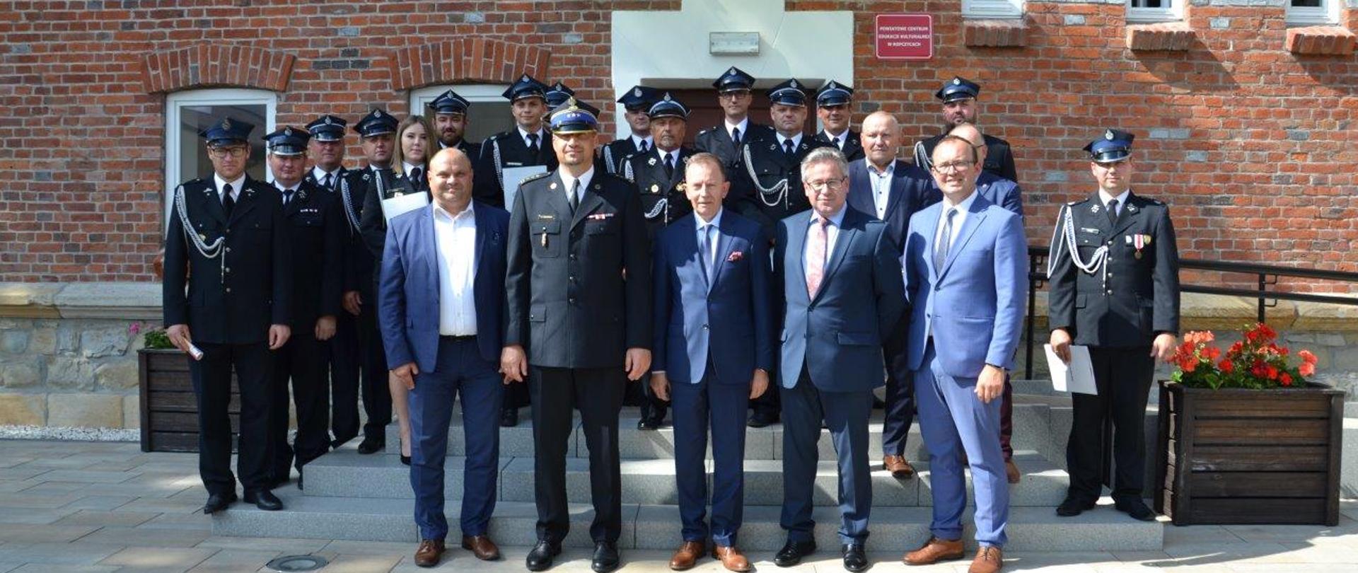 Zdjęcie przedstawia plac przed wejściem do Powiatowego Centrum Edukacji Kulturalnej w Ropczycach na którym stoją przedstawiciele jednostek OSP w umundurowaniu wyjściowym wytypowani do podpisania umów z WFOŚiGW.. Na pierwszym planie przed druhami OSP stoją od lewej: Starosta Powiatu ropczycko-sędziszowskiego Witold Darłak, Komendant Powiatowy PSP w Ropczycach st. bryg. Witold Wiercioch, Zastępca Prezesa Zarządu WFOŚiGW w Rzeszowie Bogusław Kida, Poseł RP Kazimierz Moskal oraz Wójt Gminy Ostrów Grzegorz Ożóg.