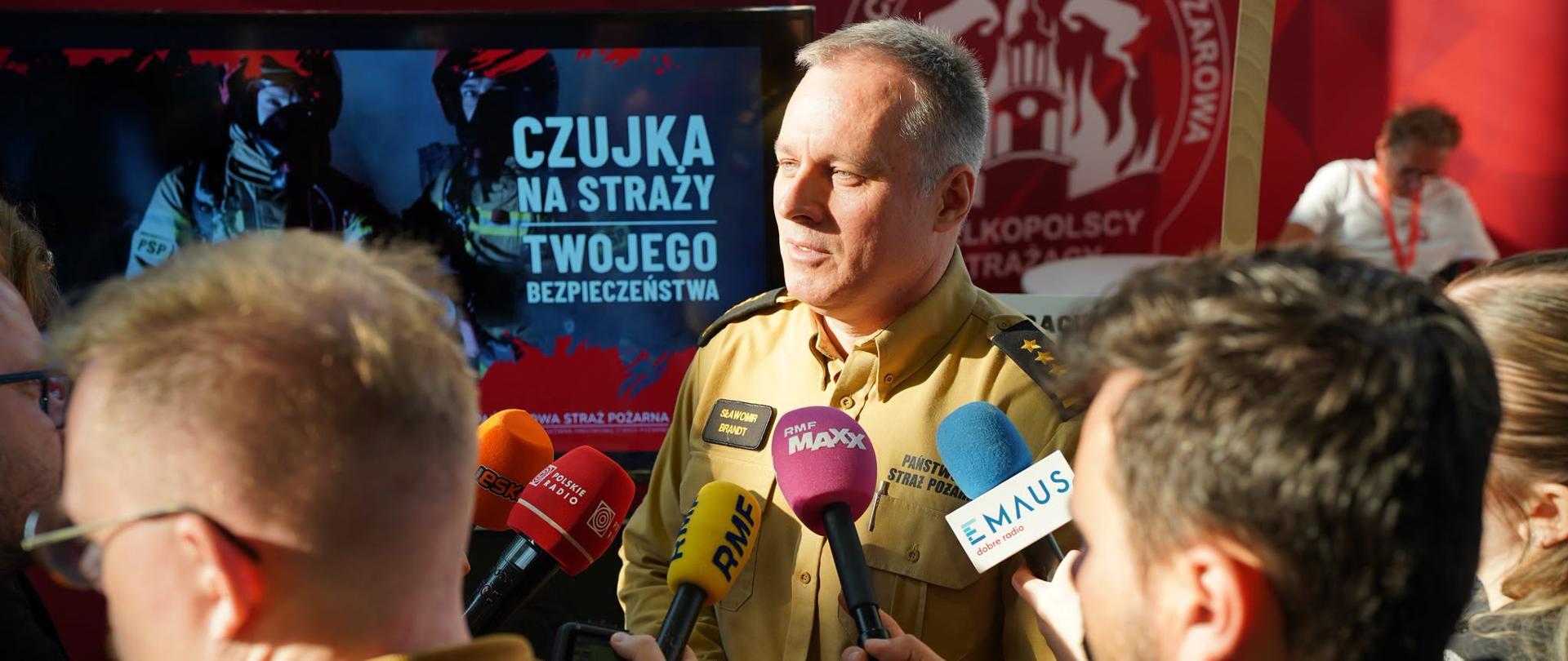 Strażak stoi i do mówi do mikrofonu wewnątrz hali wystawowej, w tle ścianka reklamowa PSP i namiot z wystawą dotyczącą ochroną ludności. W tle statystyka interwencji strażaków oraz prezentacje czujek i materiałów. 