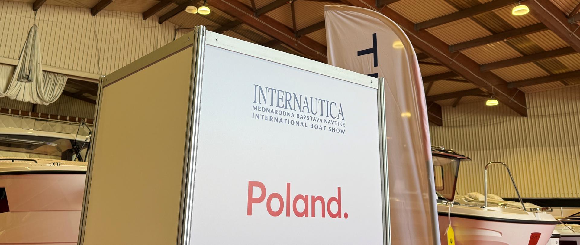 Tablica z oznaczeniem „Poland” na stoisku narodowym podczas targów Internautica, otoczona łodziami polskich producentów.