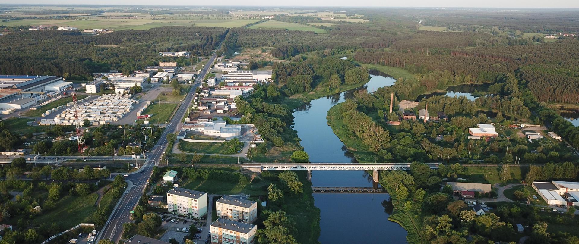 Zdjęcie przedstawia widok z lotu ptaka na krajobraz, który obejmuje mieszankę elementów naturalnych i miejskich. Widoczne są liczne budynki, niektóre z dużymi płaskimi dachami, co wskazuje na ich przemysłowe lub komercyjne zastosowanie, a inne są mieszkalne z dachami skośnymi. Rzeka meandruje przez scenę, otoczona zielenią i drzewami. Przez rzekę przechodzi most kolejowy.