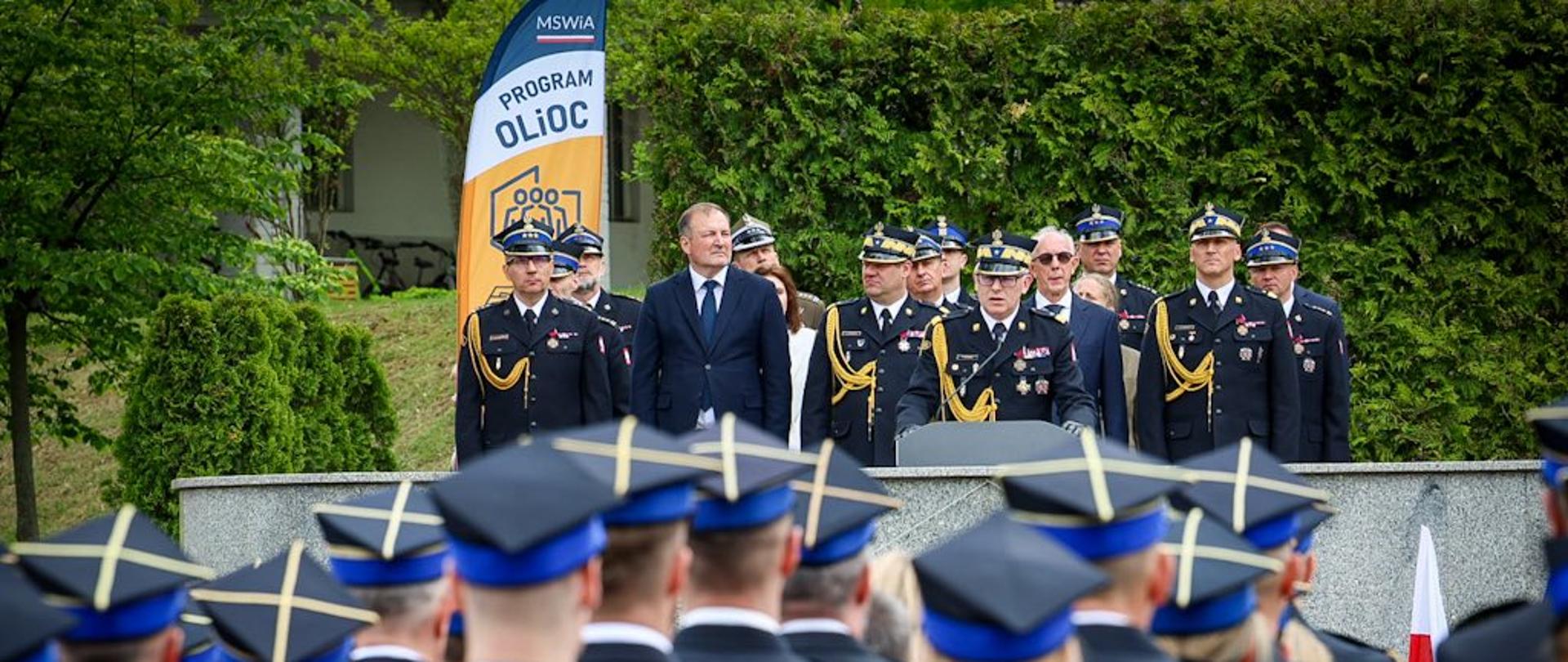  Do mikrofonu przemawia Zastępca Komendanta Głównego PSP nadbryg. Paweł Frysztak. Za nim goście uroczystości. Wśród nich Komendant Miejski PSP m. st. Warszawy st. bryg. Paweł Adamczyk, Mazowiecki Komendant Wojewódzki PSP nadbryg. Artur Gonera oraz Rektor-Komendant Akademii Pożarniczej nadbryg. Tomasz Klimczak. 