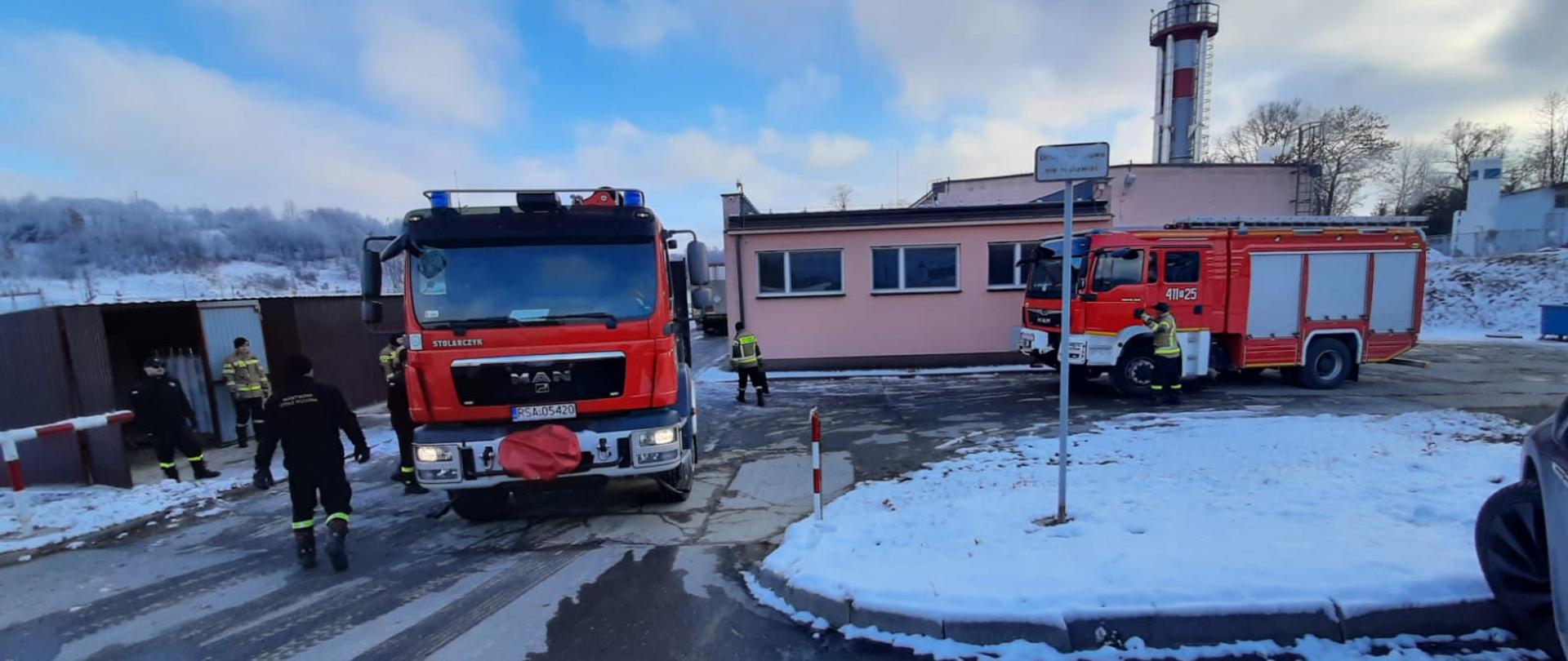 Zdjęcie zrobione na zewnątrz. Na pierwszym planie widać samochód strażacki i czterech strażaków
stojących obok samochodu. Na drugim planie w tle widać budynek i samochód strażacki, do którego
wsiada jeden strażak.