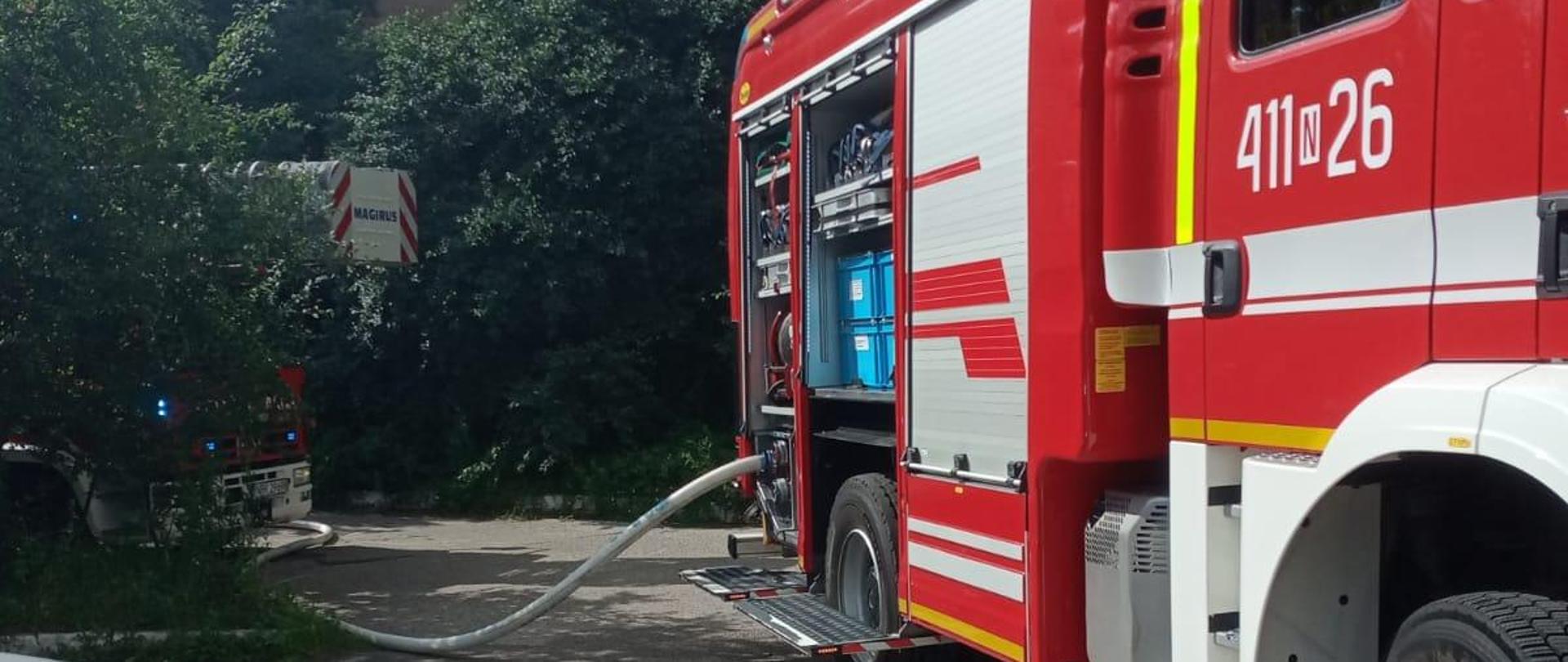 Na zdjęciu z prawej strony stoi duży samochód strażacki. W oddali zielone drzewa, krzaki. Pogodnie, słonecznie.
