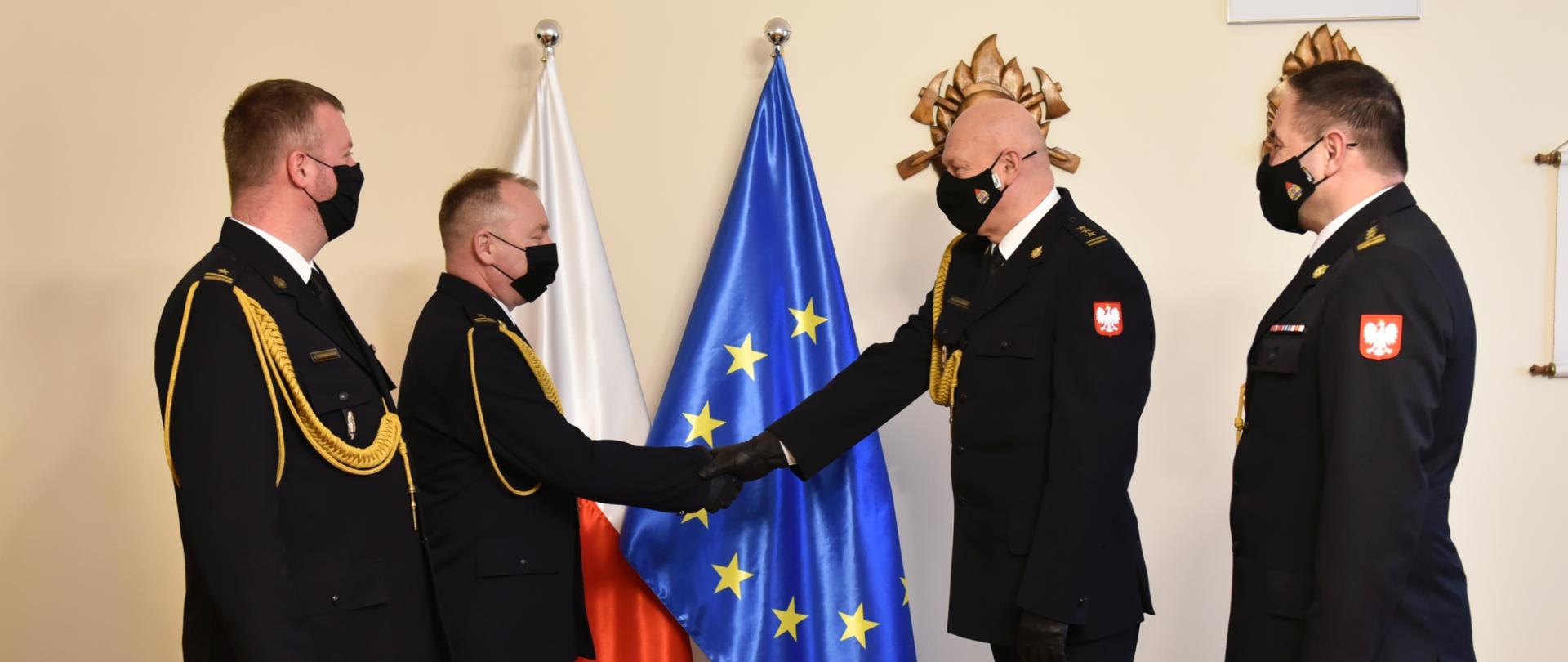 na zdjęciu widzimy od lewej Komendanta Powiatowego Państwowej Straży Pozarnej w Bełchatowie, Zastępce Komendanta Powiatowego Państwowej Straży Pozarnej w Bełchatowie, Zastępce Komendanta Wojewódzkiego Państwowej Straży Pozarnej w Łodzi, który gratuluje Zastępcy Komendana Powiatowego w Bełchatowie oraz Naczelnika wydziału kadr Komendy Wojewódzkiej Państwowej Straży Pozarnej w Łodzi, na środku flagi Polski oraz Unii europejskiej.