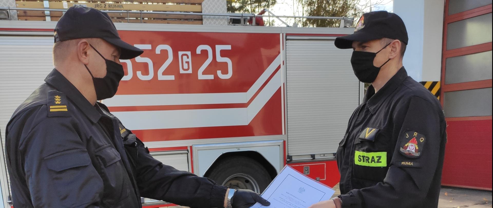 Zdjęcie przedstawia wręczenie aktu nadania wyższego stopnia służbowego przez komendanta powiatowego funkcjonariuszowi, w tle wóz strażacki.