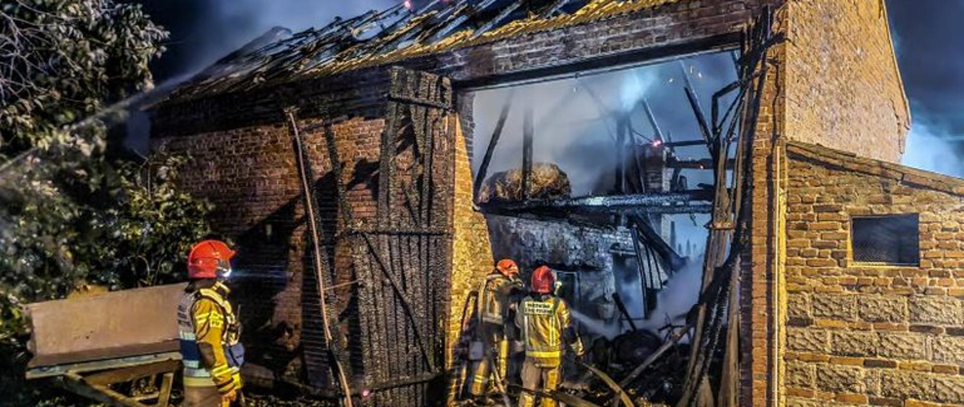 Na zdjęciu stodoła objęta pożarem. Widoczne spalone bramy wjazdowe oraz więźba dachowa. Przed budynkiem stoją strażacy, dokonując oględzin. Wewnątrz widoczne zadymienie. Pożar jest już ugaszony. 