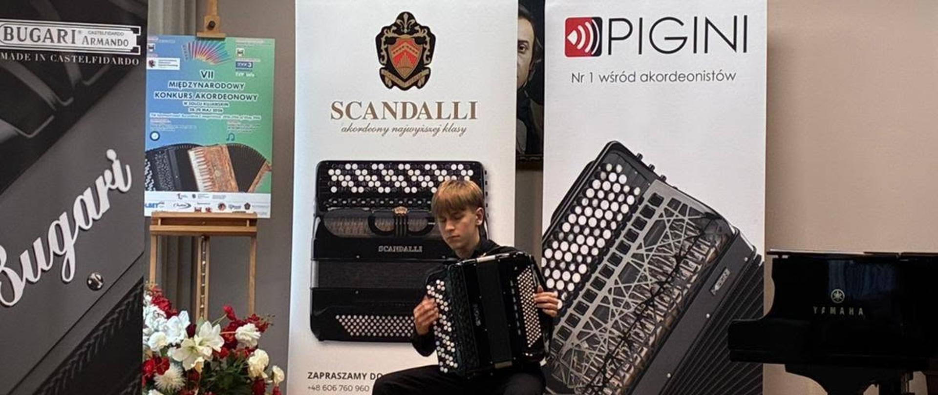 Chłopiec w czarnej koszuli siedzi na scenie i gra na czarnym akordeonie. Za nim stoją roll-upy firm Scandalli, Pigini oraz Bugari. Na scenie widoczny jest również fortepian marki Yamaha, bukiet kwiatów i godło Polski na ścianie.
