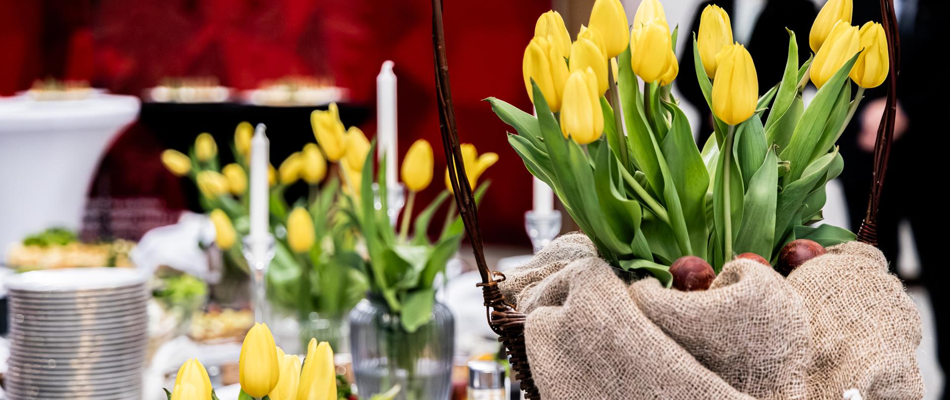 Kosz z żółtymi tulipanami ustawiony na stole cateringowym z potrawami i świecami podczas uroczystości.