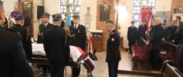 Strażacy stoją obok trumny obłożonej flagą Polski przed nią znajdują się na poduszce odznaczenia oraz komendant główny Państwowej straży Pożarnej oddaje honor w ławach stoją kobiety i mężczyźni.