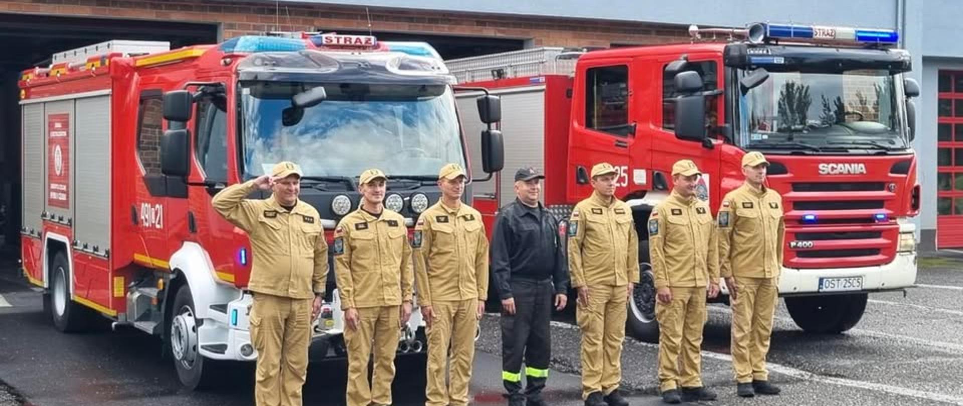 Na zdjęciu znajdują się strażacy ustawieni do zdjęcia na tle samochodów strażackich
