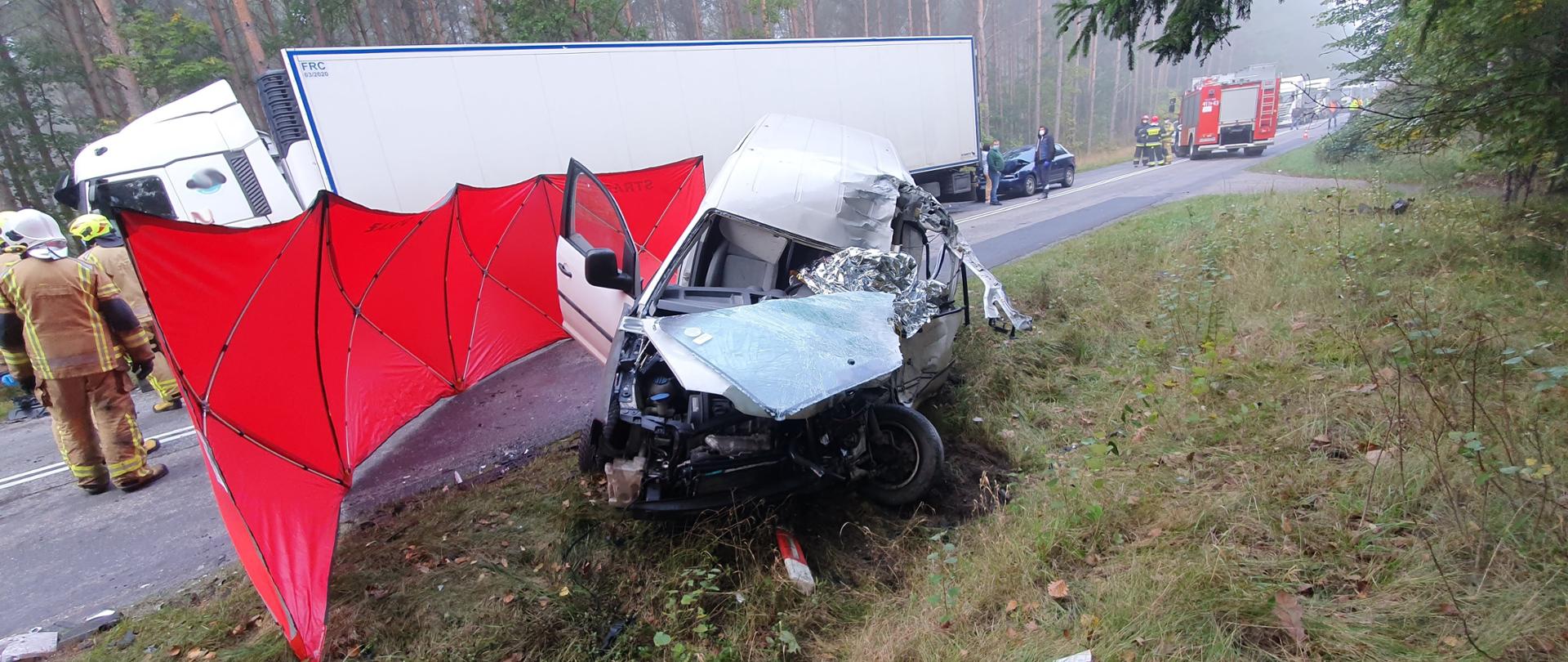 Pojazd dostawczy koloru białego znajduje się częściowo w przydrożnym rowie, Pojazd otoczony czerwonym parawanem od strony jezdni. Pojazd jest mocno rozbity, widać całkowicie popękaną przednią szybę jak i maskę i zderzak samochodu. We wnętrzu pojazdu srebrna folia zasłaniająca zwłoki ofiary śmiertelnej tego wypadku. W tle zdjęcia widać samochód ciężarowy z naczepą koloru białego i grupę 3 strażaków ubranych w ubrania specjalne. 