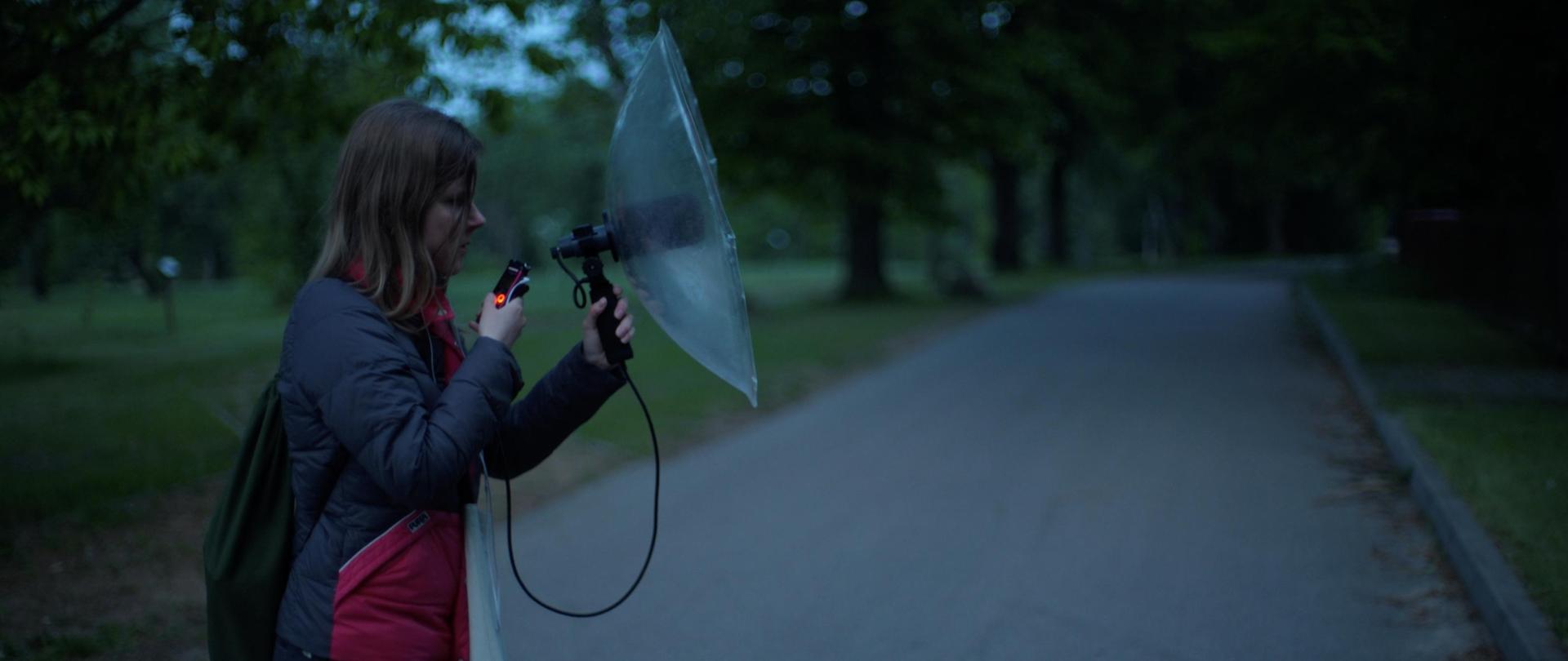 Izabela Dłużyk is standing with a microphone in her left hand and a remote control in her right, recording. It's dark and gray outside, around dawn.