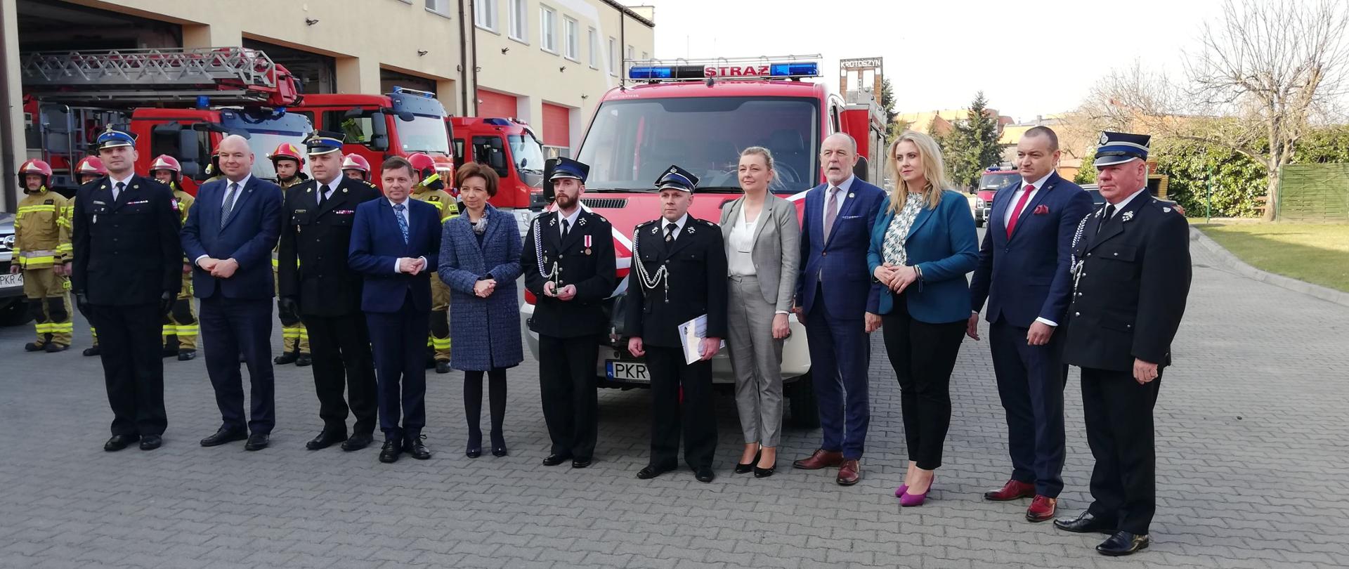 na dużym placu grupa ludzi stoi przed czerwonym autem strażackim