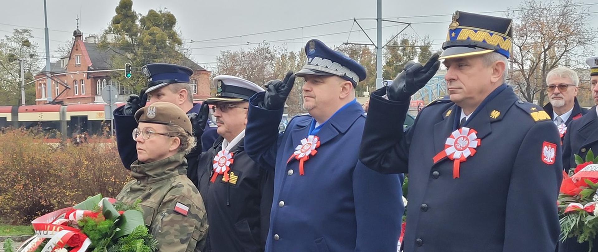 Przedstawiciele służb mundurowych podległych Ministerstwu Spraw Wewnętrznych i Administracji stoją obok siebie oddając honory przed nimi stoi żołnierz który trzyma wieniec.