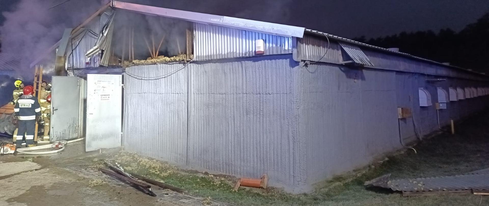 Zdjęcie przedstawia budynek kurnika gdzie doszło do pożaru. Z lewej strony pracują strażacy.