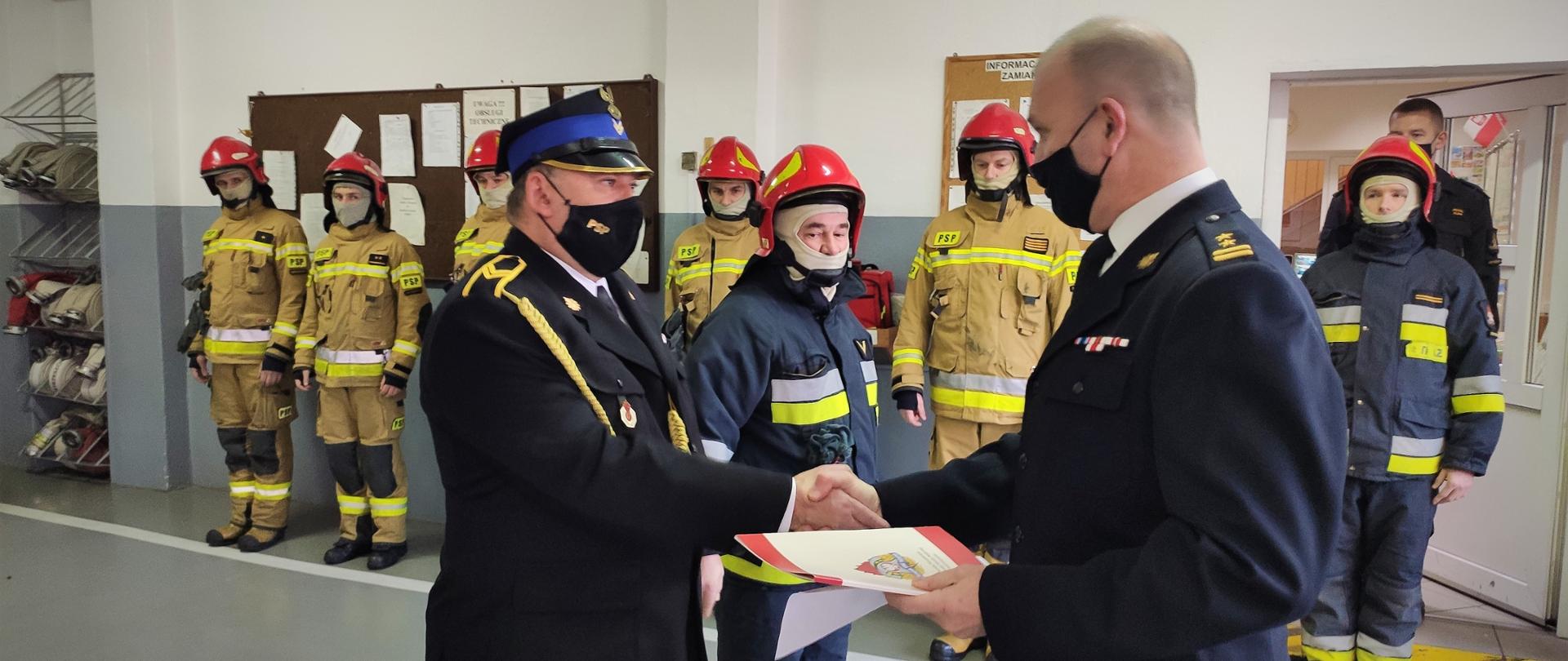 Na pierwszym planie Komendant Powiatowy Państwowej Straży Pożarnej w Ostrzeszowie w trakcie uścisku dłoni z jednym z funkcjonariuszy, którzy odchodzą na prawa emerytalne. Obok stoi drugi funkcjonariusz. W tle stoją strażacy w umundurowaniu specjalnym podczas wydarzenia.