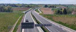 Widok z góry na dwujezdniową drogę, dookoła pola uprawne, nieliczne drzewa i zabudowania, pogodne niebo