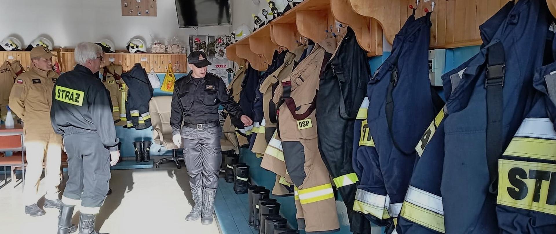 Zdjęcie przedstawia wnętrze remizy Ochotniczej Straży Pożarnej w Piekoszowie. Goście zwiedzają szatnię gdzie druhowie przechowują umundurowanie bojowe.