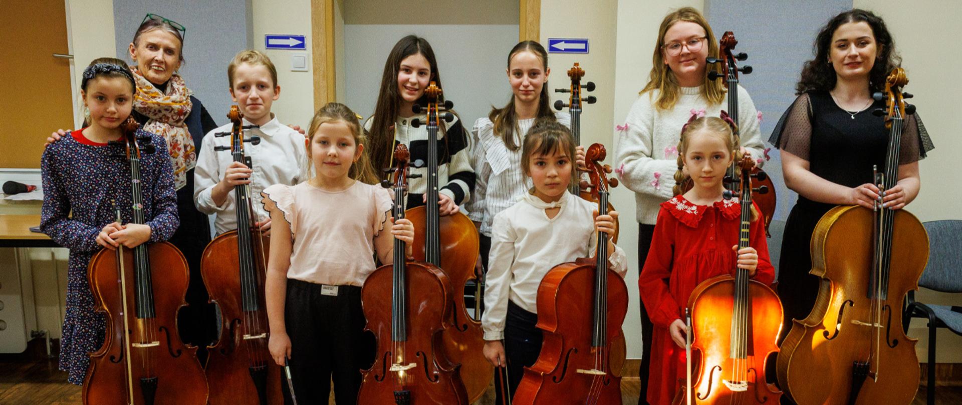 Dwie nauczycielki i kilkoro uczniów stoją w auli szkolnej, w rękach trzymają wiolonczele.