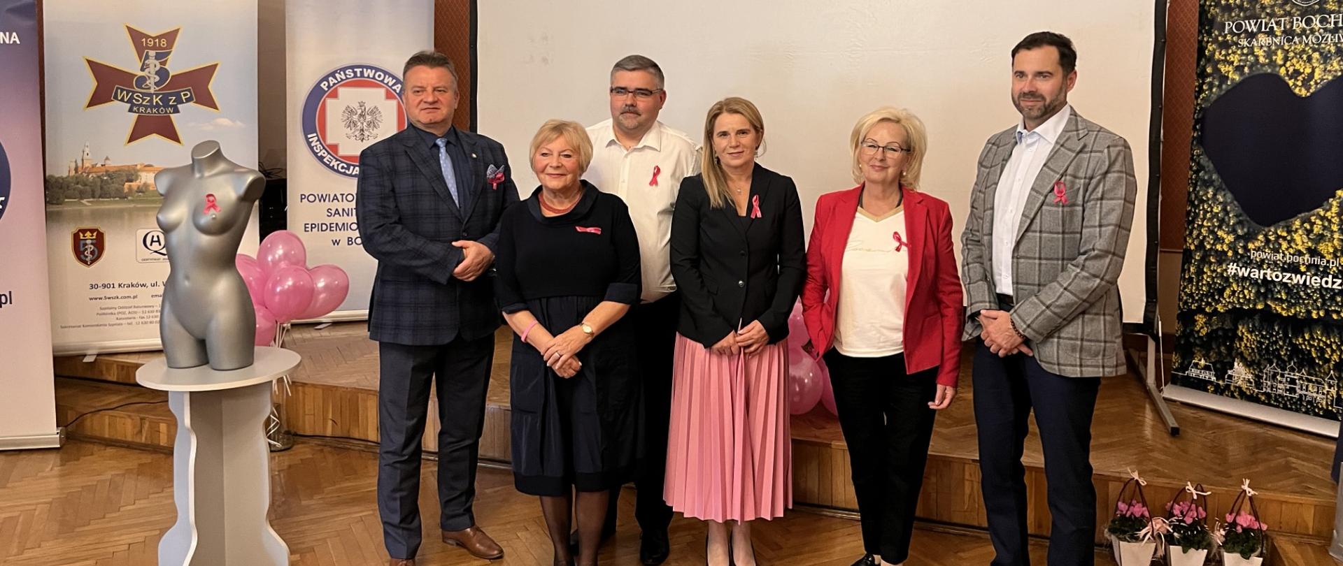 W tle znajduje się ekran. Przed ekranem stoją mężczyźni i kobiety. Po lewej popiersie kobiety.