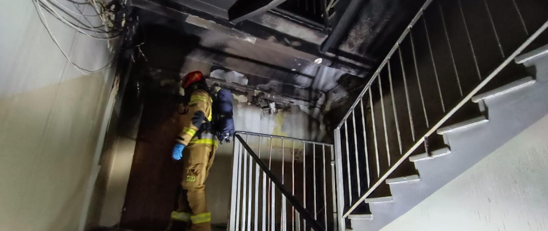 Strażak w pełnym umundurowaniu i aparacie ochrony układu oddechowego dokonuje inspekcji po pożarze na klatce schodowej.
