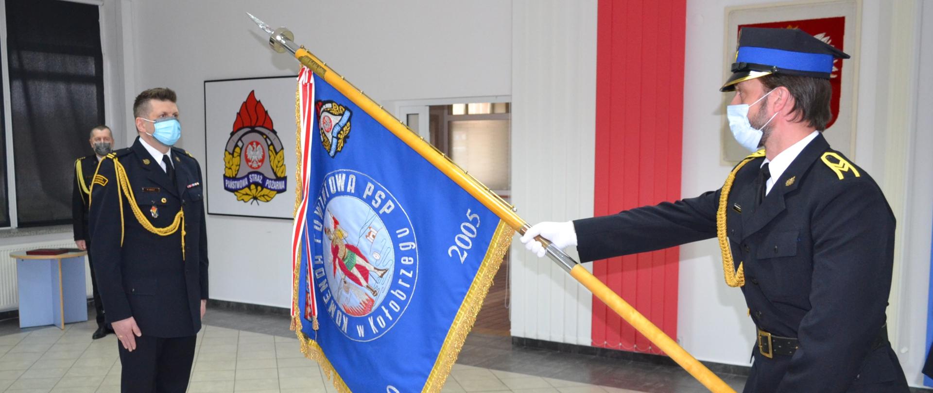 Strażak w maseczce, stojący na baczność, naprzeciw niego strażak pochylający sztandar komendy