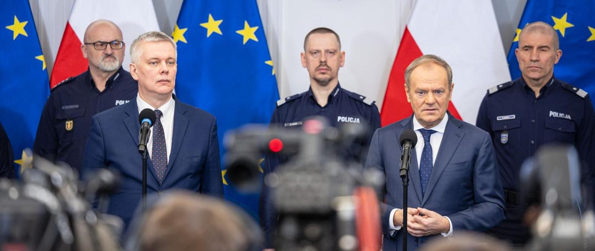  Dwaj mężczyźni w garniturach, oraz trzej w policyjnych mundurach na tle flag Polski i Unii Europejskiej stojący przed kamerami na konferencji prasowej