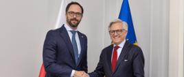 Dwóch mężczyzn wymienia uścisk dłoni. Są formalnie ubrani i uśmiechają się.