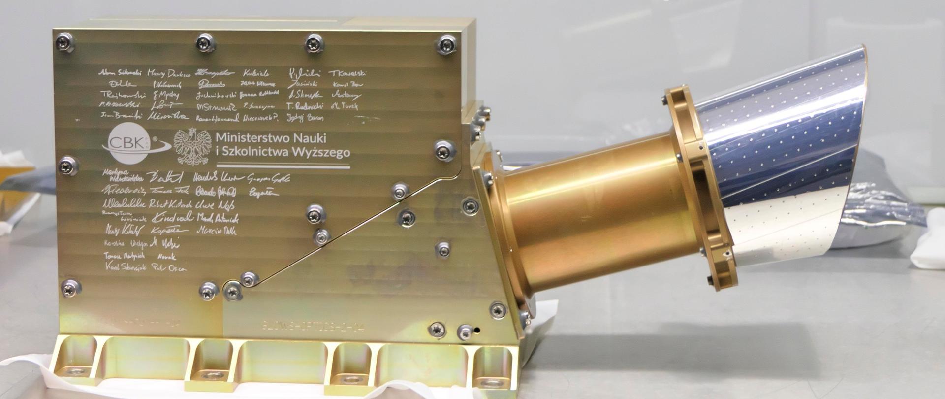 Zdjęcie urządzenia - złociste prostokątne pudełko z wystającą z prawej złoto - srebrną tubą.