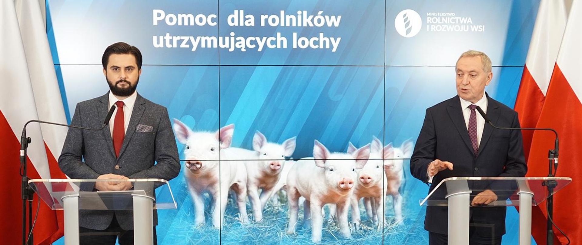 Konferencja prasowa Pomoc dla producentów trzody chlewnej. Na tle prezentacji dwie mównice. Przy jednej z nich stoi minister Henryk Kowalczyk, przy drugiej sekretarz stanu - Norbert Kaczmarczyk.