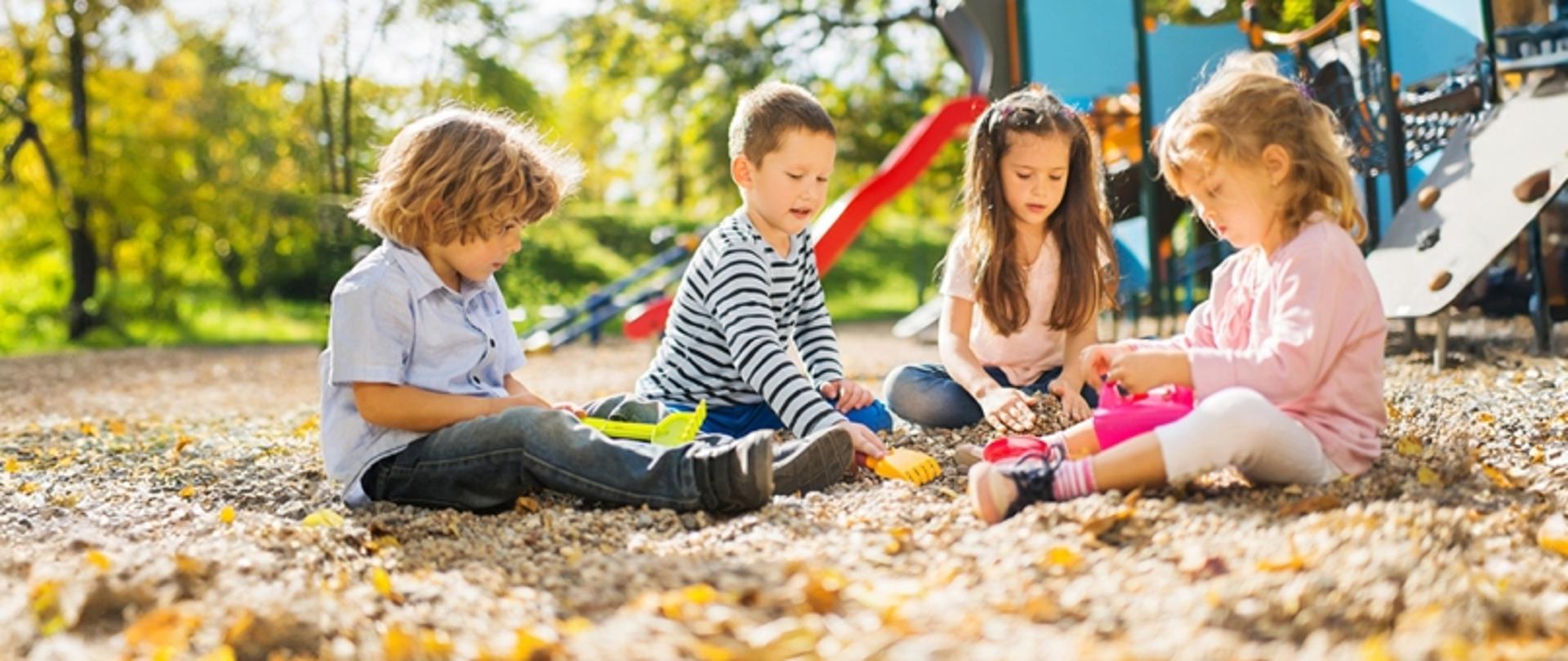 Na placu zabaw czworo dzieci bawi się siedząc na piachu. W tle ścianka wspinaczkowa i zjeżdżalnia.