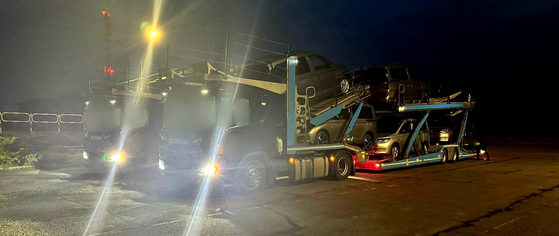 Nocna scena na punkcie kontrolnym: ciężarówka typu laweta przewożąca kilka samochodów osobowych stoi przy krawężniku, oświetlona jasnym światłem latarni. W tle ciemność i lekka mgła, obok chodnik i znak drogowy zakazu.