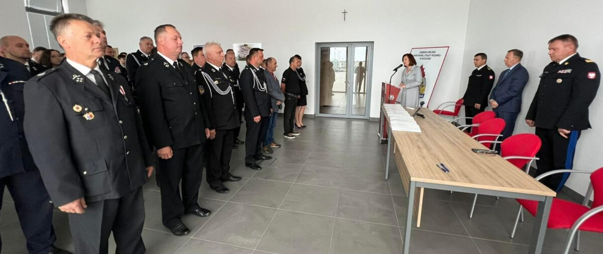 Zdjęcie zrobione w pomieszczeniu, w sali konferencyjnej. Po lewej stronie ustawieni strażacy Ochotniczych Straży Pożarnych w mundurach wyjściowych. Po prawej stronie, obok stołu prezydialnego stoją cztery osoby, wśród nich dwóch oficerów Państwowej Straży Pożarnej w mundurach wyjściowych. 