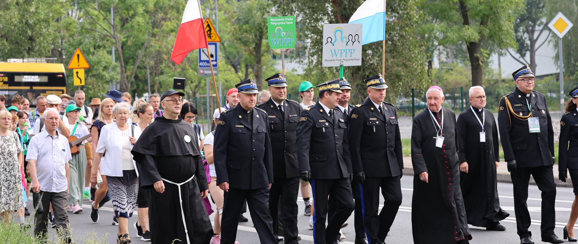 Grupa uczestników Warszawsko-Praskiej Pielgrzymki Pieszej (WPPP) idąca miejską ulicą. Na czele pochodem kroczą biskup, księża, zakonnik w habitach oraz strażacy w galowych mundurach. Uczestnicy niosą polską flagę, biało-niebieską flagę maryjną oraz tablice z logo pielgrzymki. W tle widać miejskie drzewa, znaki drogowe oraz budynek biurowy.