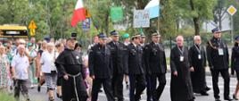 Grupa uczestników Warszawsko-Praskiej Pielgrzymki Pieszej (WPPP) idąca miejską ulicą. Na czele pochodem kroczą biskup, księża, zakonnik w habitach oraz strażacy w galowych mundurach. Uczestnicy niosą polską flagę, biało-niebieską flagę maryjną oraz tablice z logo pielgrzymki. W tle widać miejskie drzewa, znaki drogowe oraz budynek biurowy.