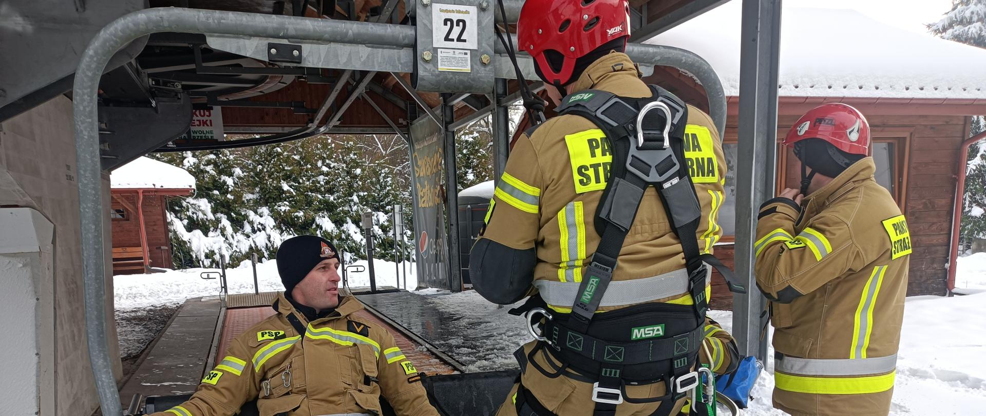 Na zdjęciu na pierwszym planie widać czterech strażaków. Dwóch z nich siedzi na wyciągu krzesełkowym przy stacji narciarskiej - jako poszkodowani (pozoranci). Trzeci ratownik ubrany w sprzęt do wysokościówki zwisa nad poszkodowanym. Czwarty ubiera kask zabezpieczający. Zdjęcie wykonane w scenerii śnieżnej przy stacji narciarskiej w Bałtowie.