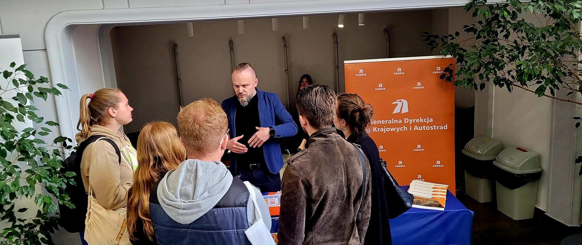 Zdjęcie przedstawia stoisko Generalnej Dyrekcji Dróg Krajowych i Autostrad (GDDKiA) na targach pracy organizowanych przez Politechnikę Śląską. Przedstawiciel instytucji prowadzi rozmowę z grupą zainteresowanych osób, które prawdopodobnie poszukują ofert pracy lub staży. Na stole rozmieszczone są materiały informacyjne, a w tle widoczny jest duży baner z logo GDDKiA oraz pełną nazwą organizacji. Dookoła stoiska znajdują się rośliny doniczkowe co nadaje przestrzeni bardziej uporządkowany i przyjazny charakter.